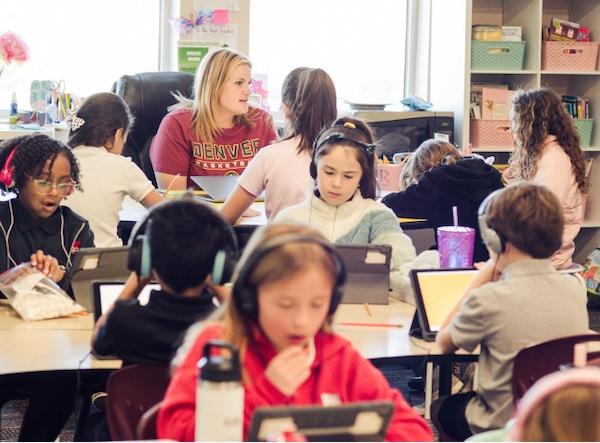 PPA elementary students are seated in a classroom learning literacy and math in their one-to-one technology support environment while a teacher works directly with four students.
